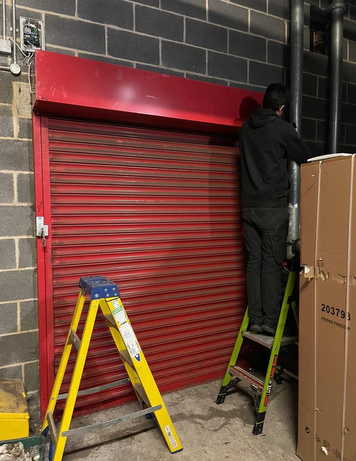 Engineer repairing a red roller shutter door from a step ladder